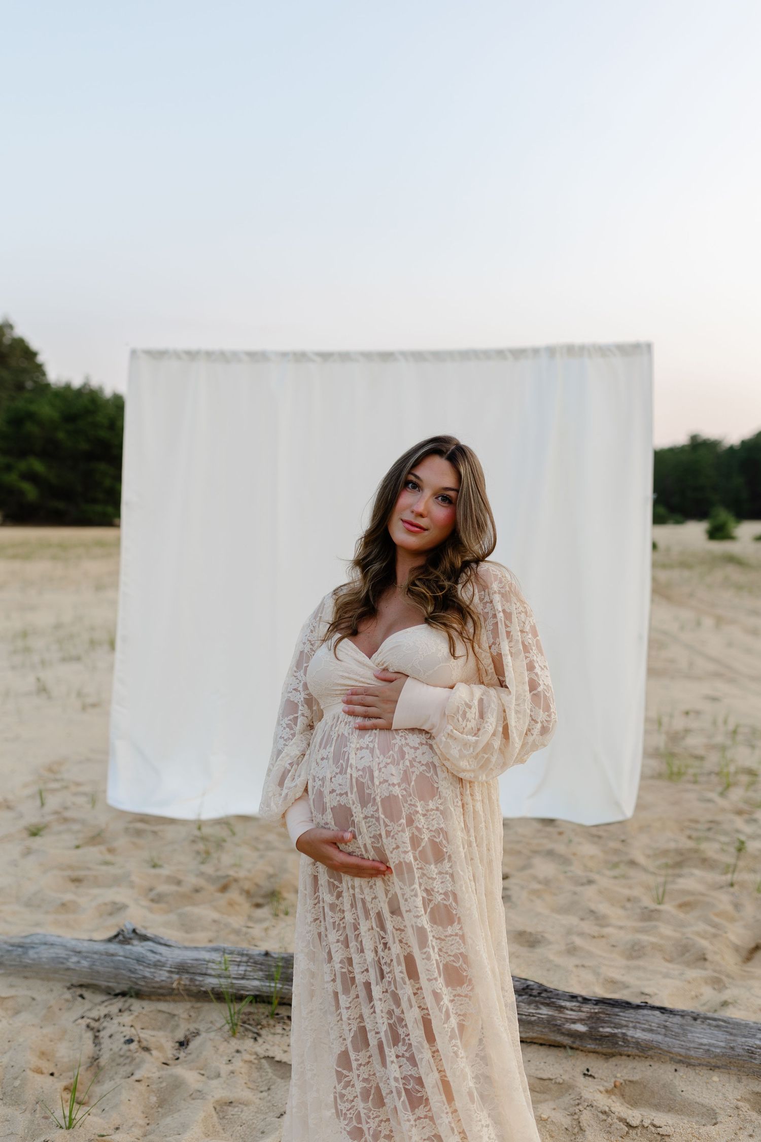 Pregnant woman in cream lace maternity gown cradles bump before white backdrop on sandy beach at dusk.