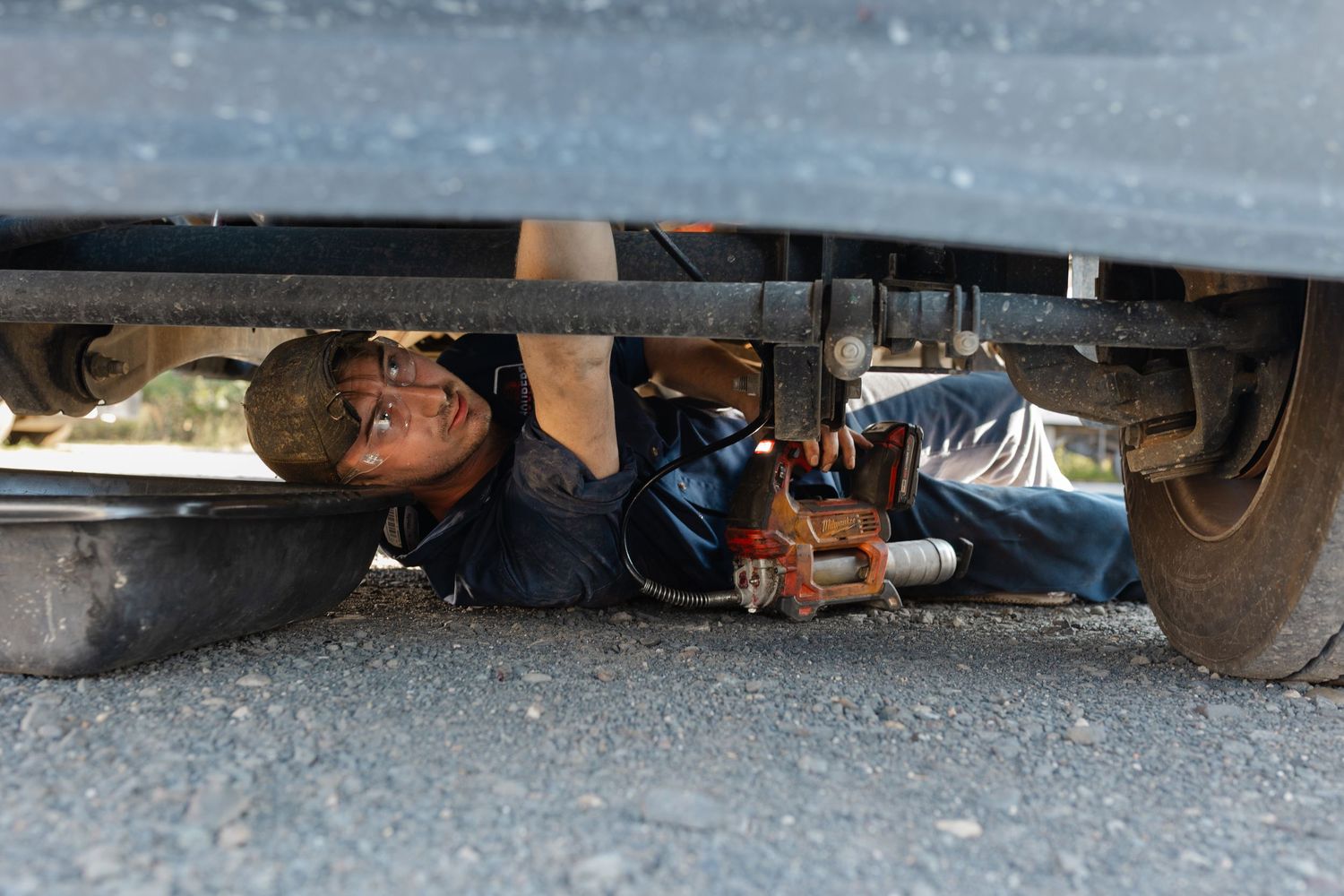 Mechanic lying under a vehicle on asphalt, using tools to repair the undercarriage of a car outdoors.