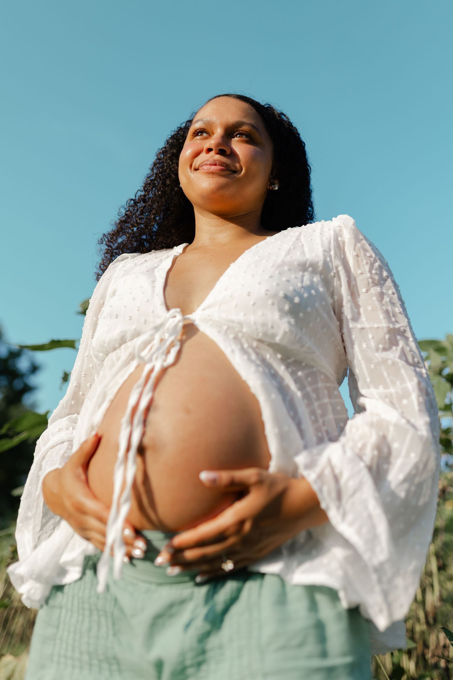 Happy pregnant woman in white blouse cradling bare baby bump under blue sky.