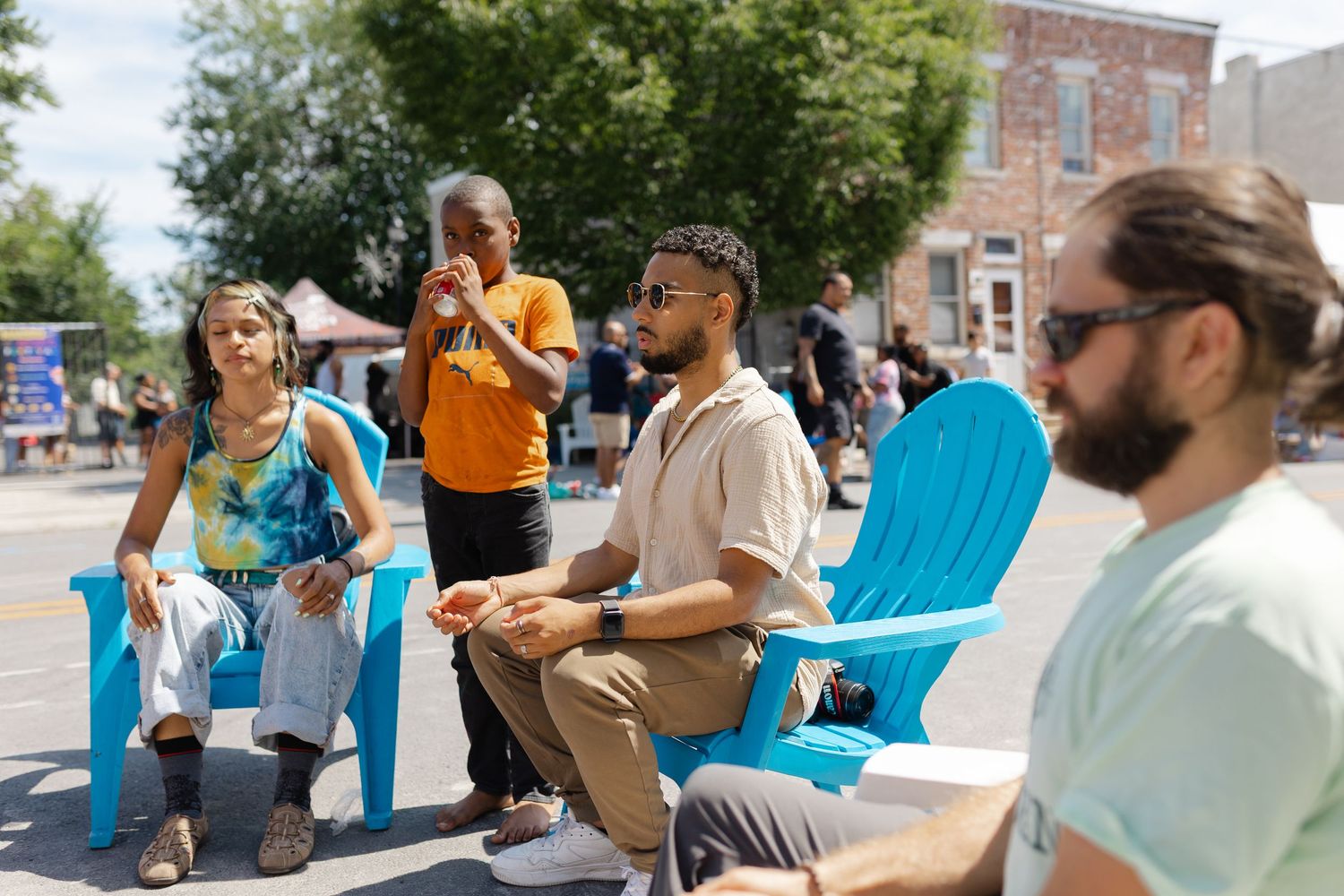 People sitting in blue Adirondack chairs outdoors on a sunny day at a street event.