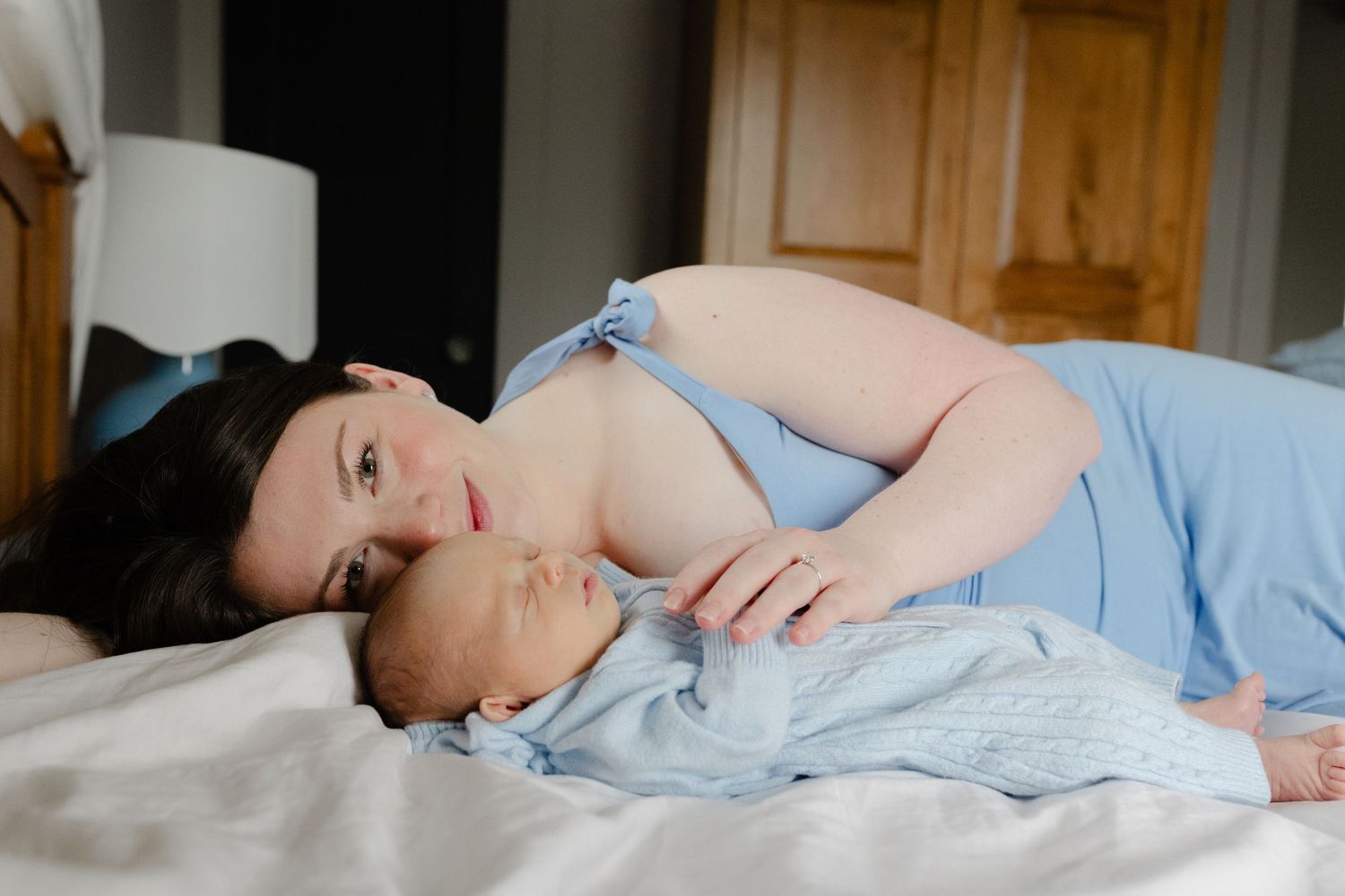 New mother in blue dress lying on bed gazing lovingly at her sleeping newborn baby.