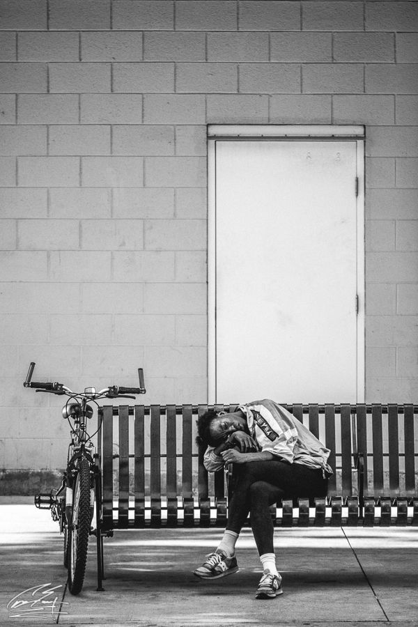 Black and white photo of a tired person sleeping on a bench next to a bicycle against a brick wall.