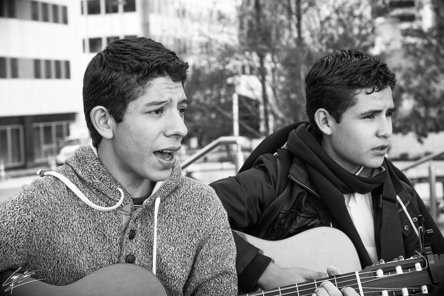 Two young musicians performing with guitars outdoors, captured candidly in a black and white urban setting.
