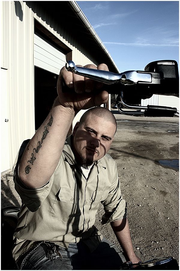 A tattooed mechanic holds up a chrome car part outside a garage under a blue sky.