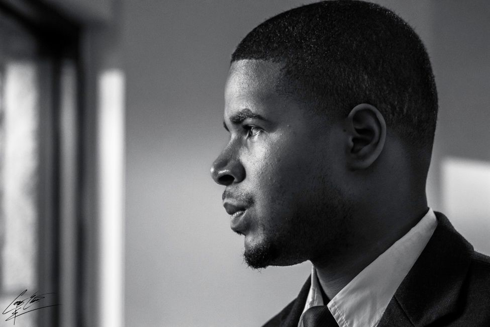 A black and white portrait of a man in profile looking out a window, with soft light illuminating his face.