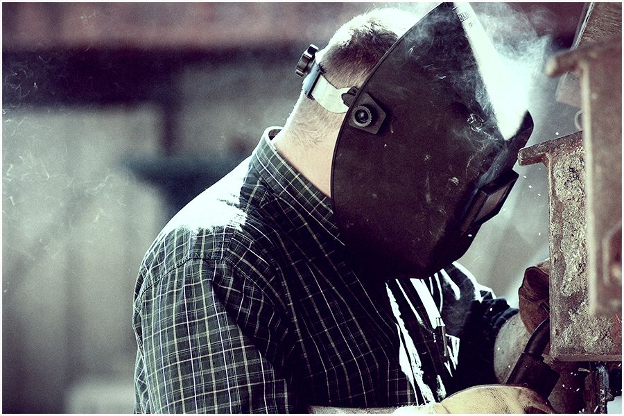 A welder wearing a protective helmet works with metal, smoke rising in an industrial workshop setting.