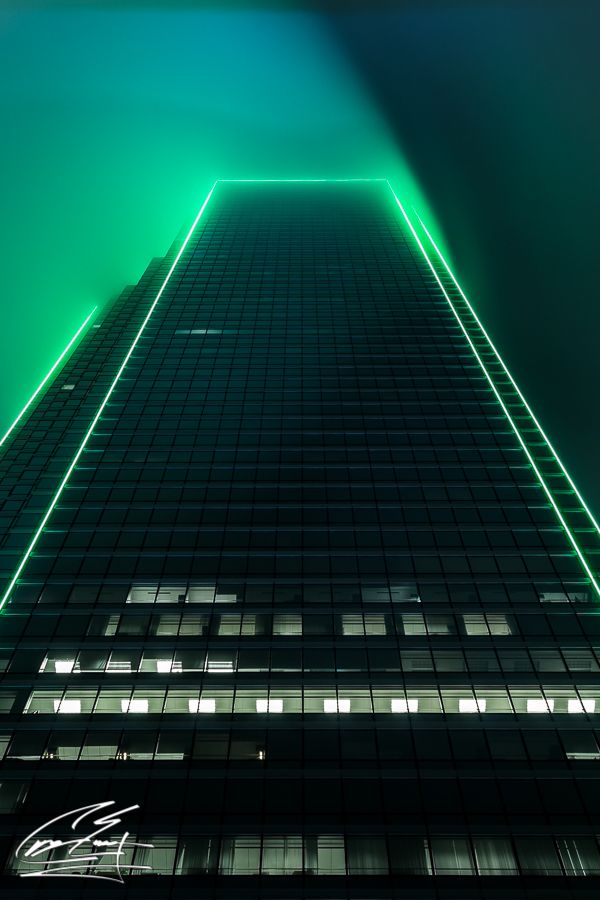 A skyscraper illuminated with vibrant green neon lights against a foggy night sky, viewed from below.