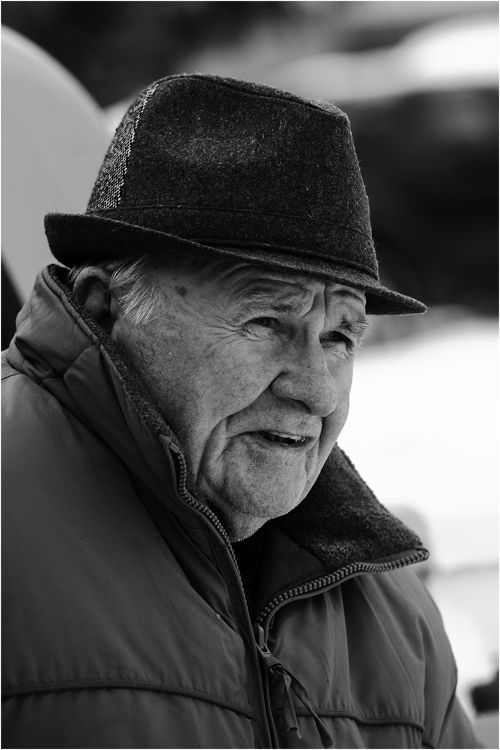 Black and white portrait of an elderly man wearing a fedora hat and puffer jacket outdoors.