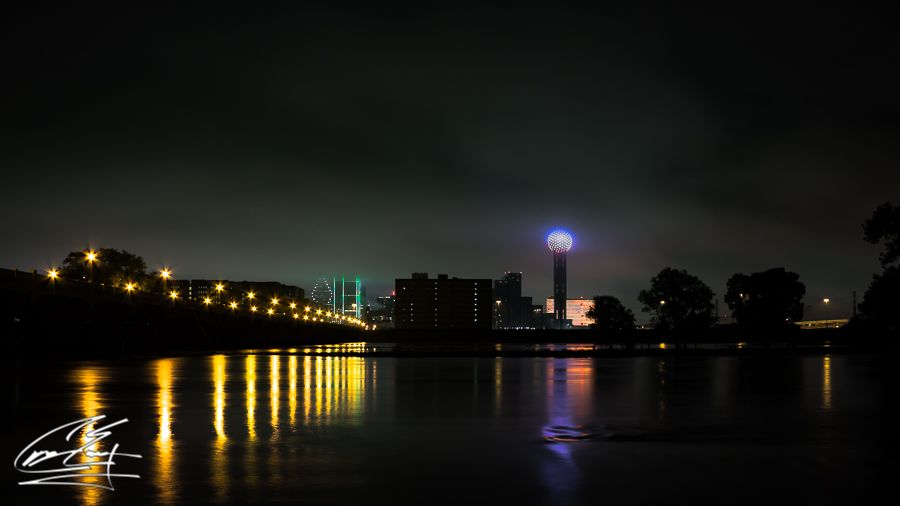 A city skyline at night with colorful lights reflecting on a calm river, featuring a prominent tower.