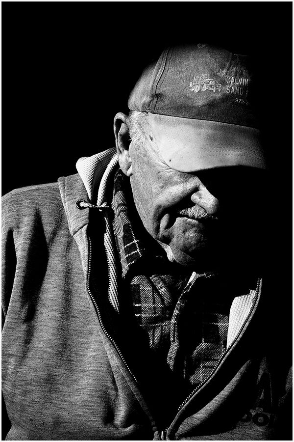 Black and white close-up portrait of an older man wearing a cap, looking downward thoughtfully.