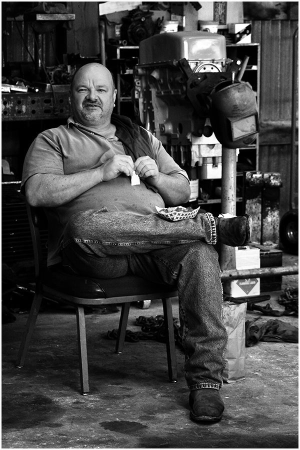 Black and white portrait of a heavyset bald man sitting in a chair inside a cluttered workshop.