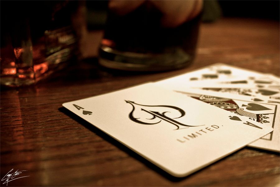 A close-up of playing cards on a wooden table beside a glass of whiskey, lit with warm ambient lighting.