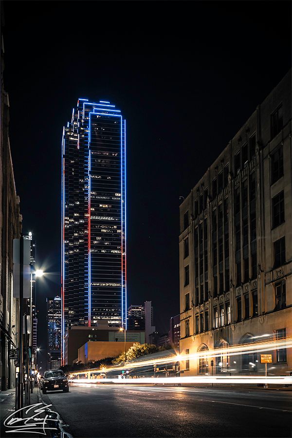 A towering skyscraper outlined in blue and red neon lights dominates a Dallas city street at night.
