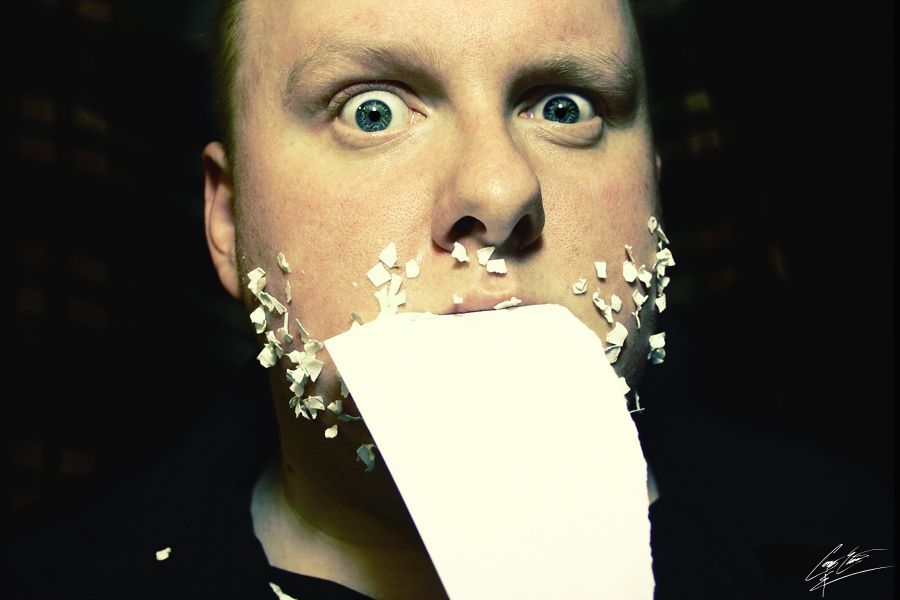 A man with wide eyes has a white card taped over his mouth with confetti scattered around his face.
