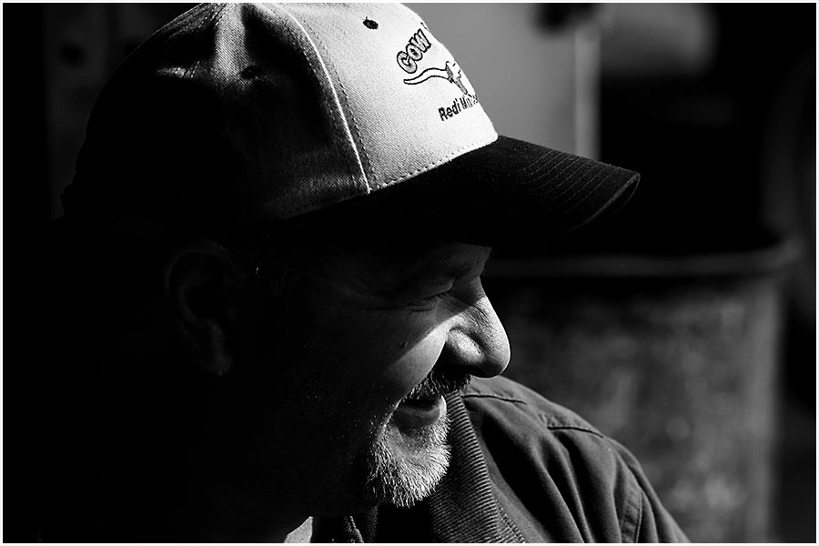 Black and white portrait of a bearded man wearing a baseball cap, smiling in profile view.
