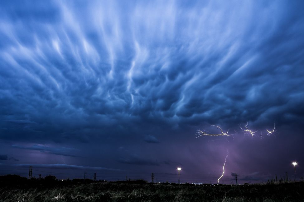 Dramatic lightning storm with mammatus clouds illuminated in deep blue and purple tones over a dark landscape.