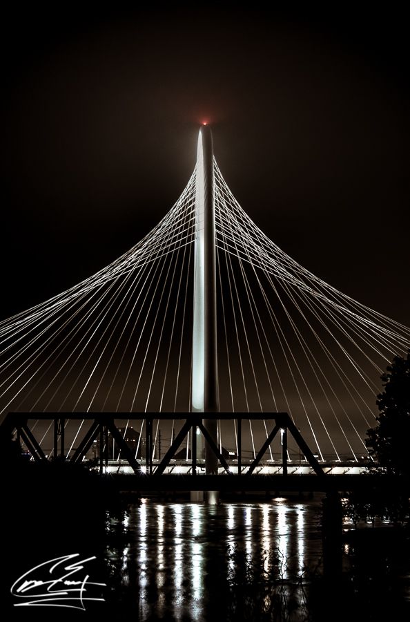 A cable-stayed bridge illuminated at night with a red light atop its pylon reflecting on calm water below.