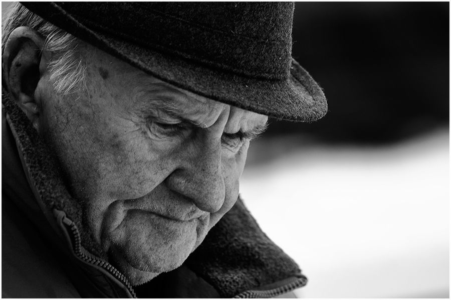 Close-up black and white portrait of a pensive elderly man in a hat looking downward.