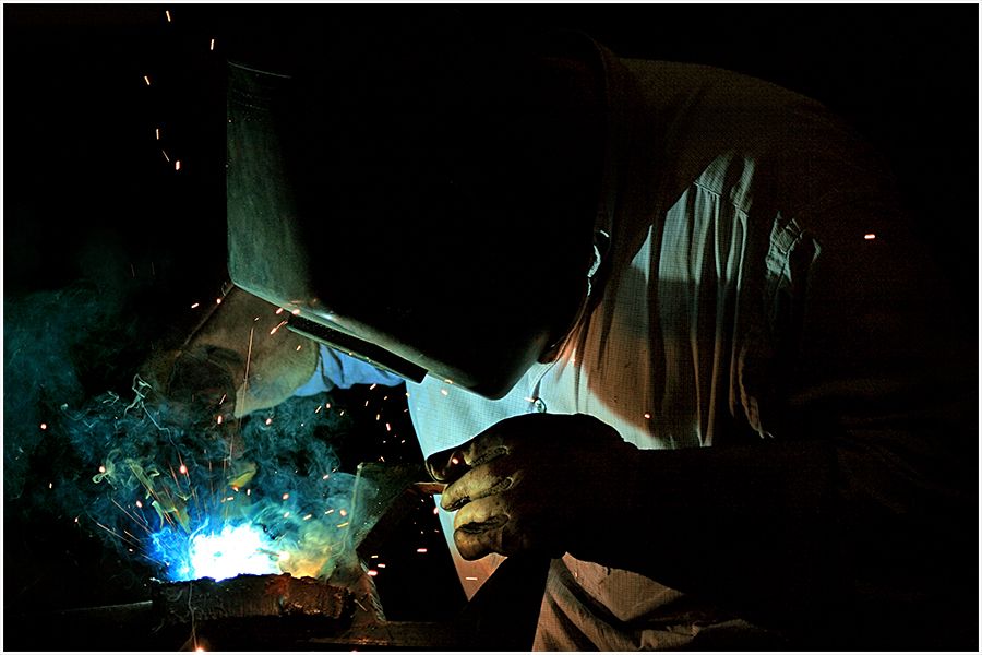 A welder works in darkness with bright blue sparks flying from the welding torch at night.