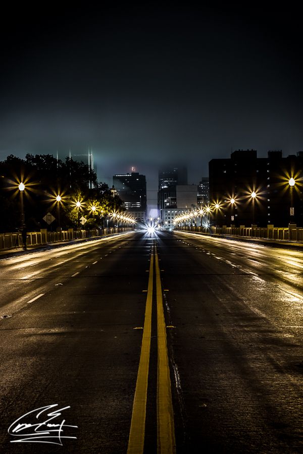 Empty wet road at night leading toward foggy city skyline with glowing street lamps.
