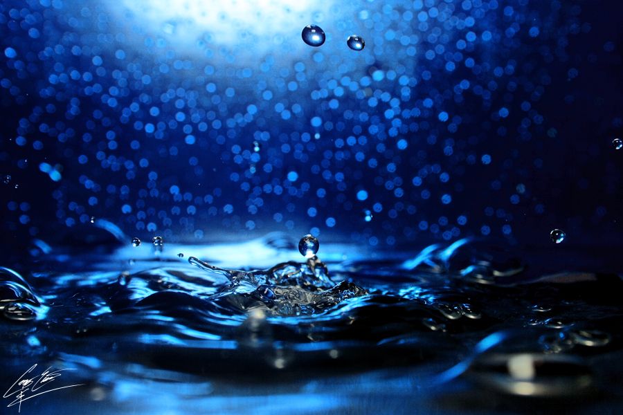 A high-speed macro photograph of a water droplet impacting a surface, creating a crown splash against a blue background.
