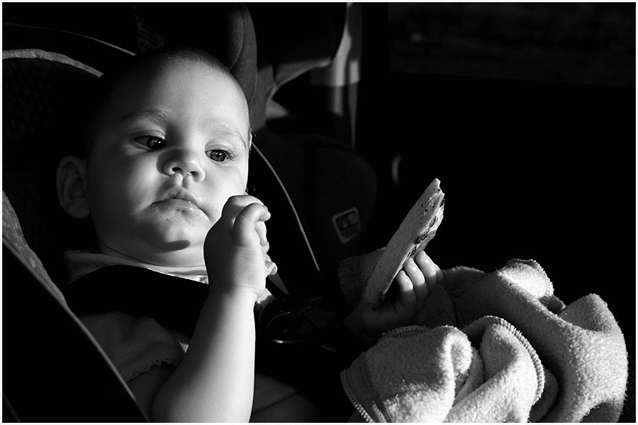 Black and white photo of a young toddler sitting in a car seat holding a stuffed animal toy.
