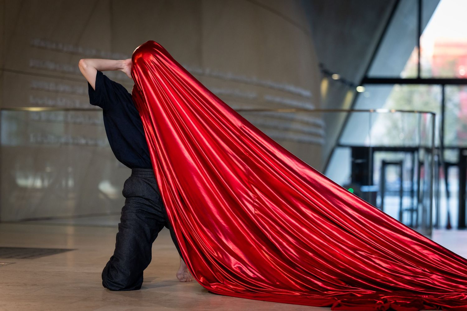 Person in black outfit dramatically holding a large shiny red fabric cape indoors.
