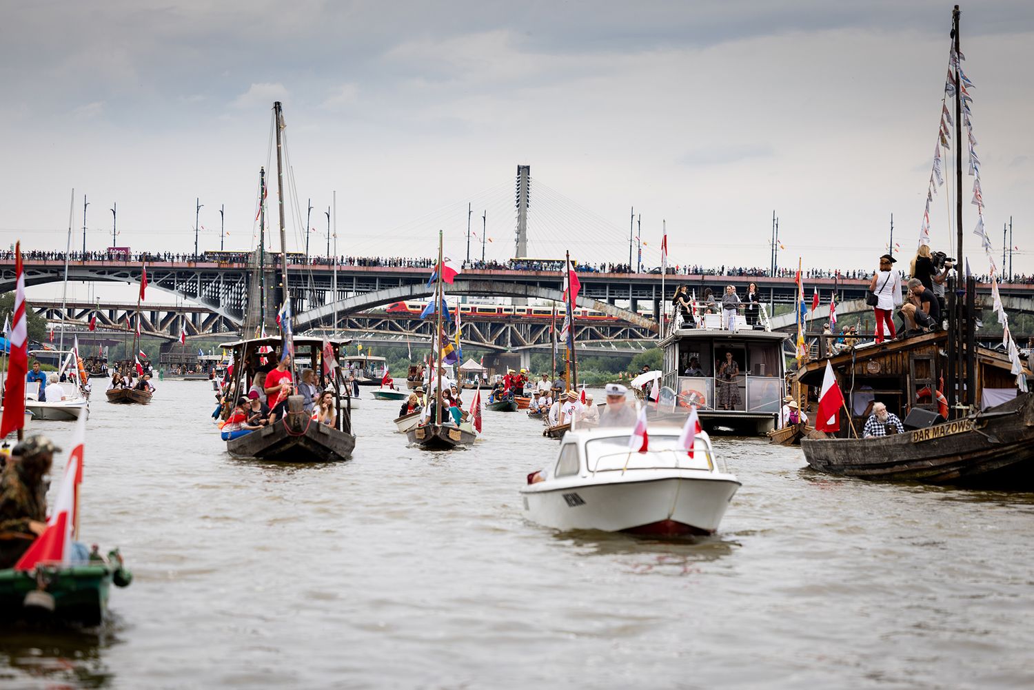 Festive boat parade on a river with colorful flags and vessels near a bridge.