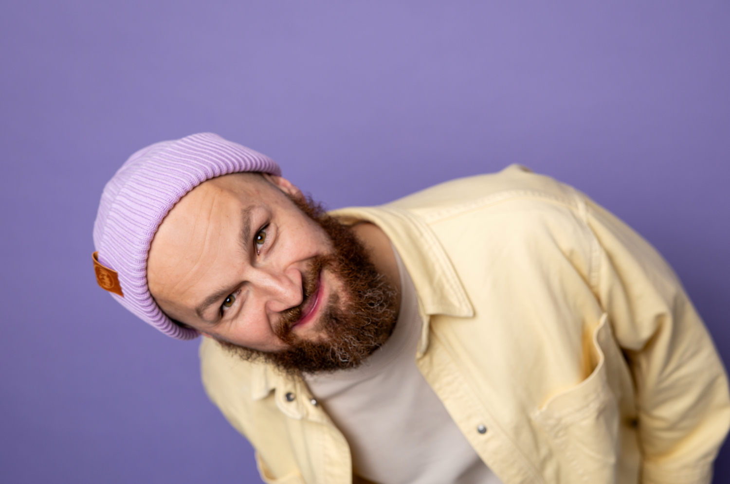 Bearded man in yellow shirt and pink beanie smiling on purple background.