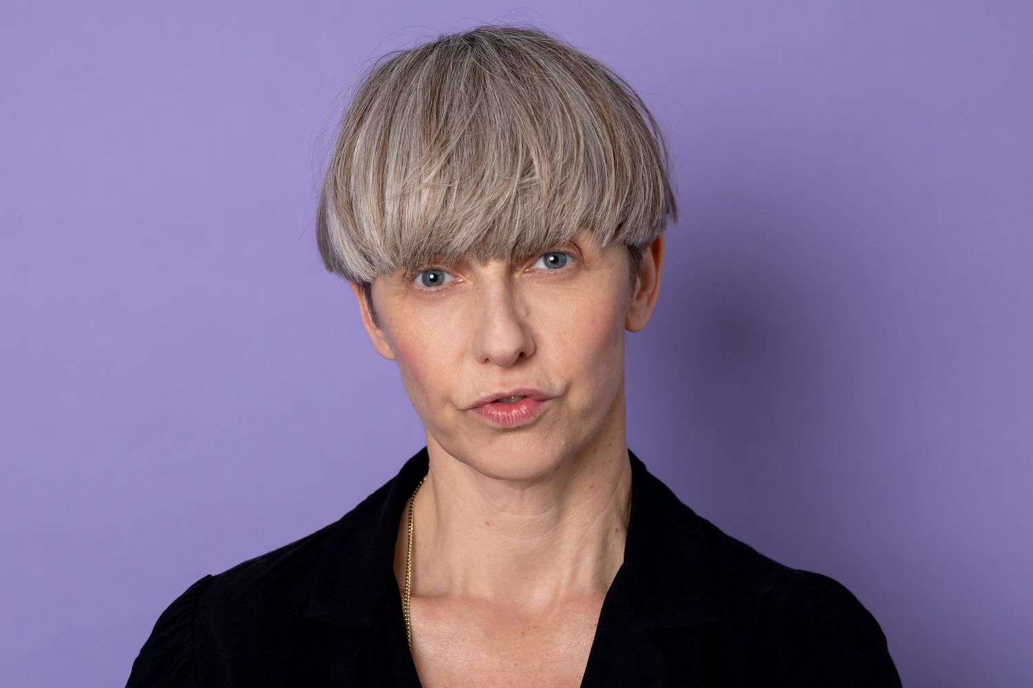 Person with short gray hair in black jacket on purple background.