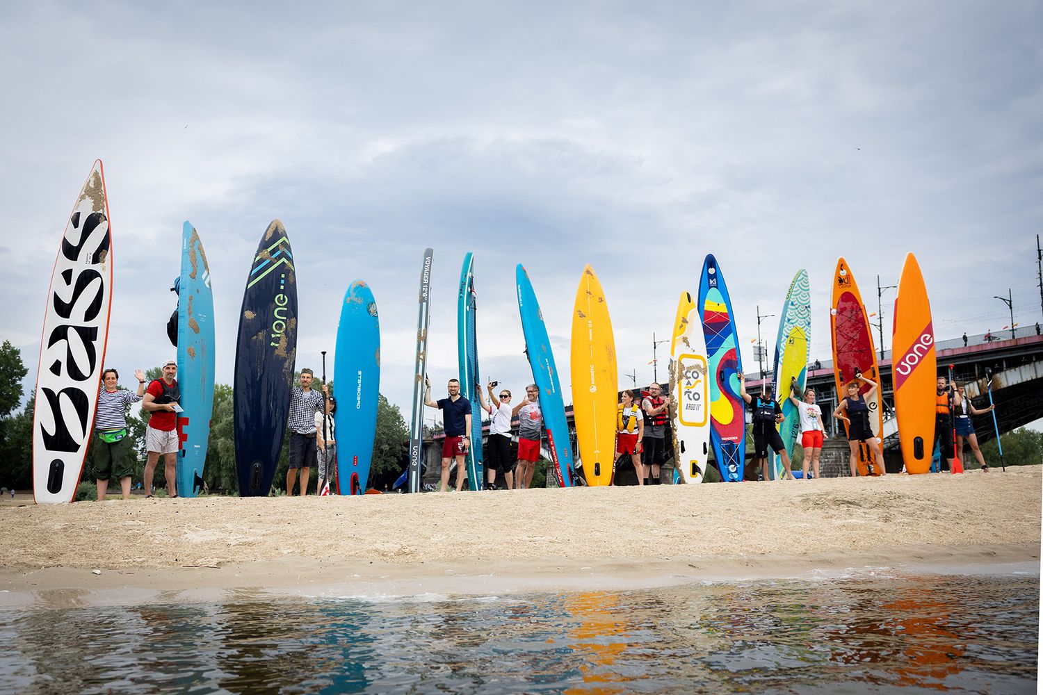 Group of people with colorful paddleboards lined up on sandy riverbank.