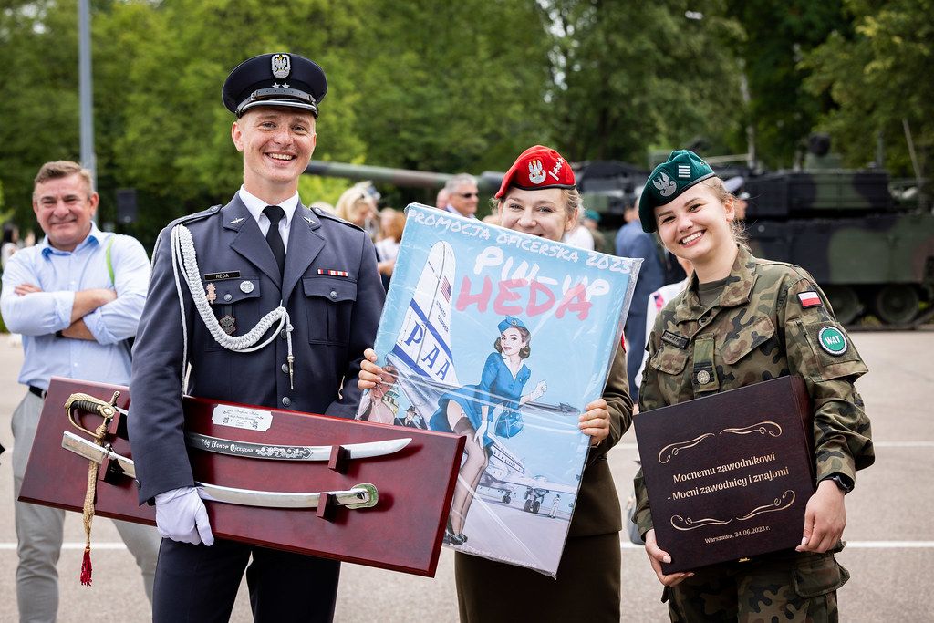 Military graduates celebrating with poster and awards at outdoor ceremony.