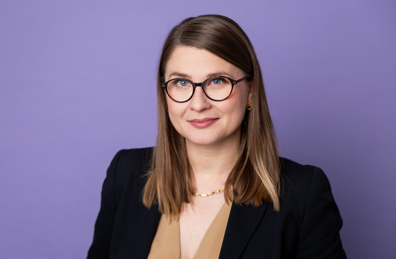 Woman with glasses and brown hair in blazer on purple background.