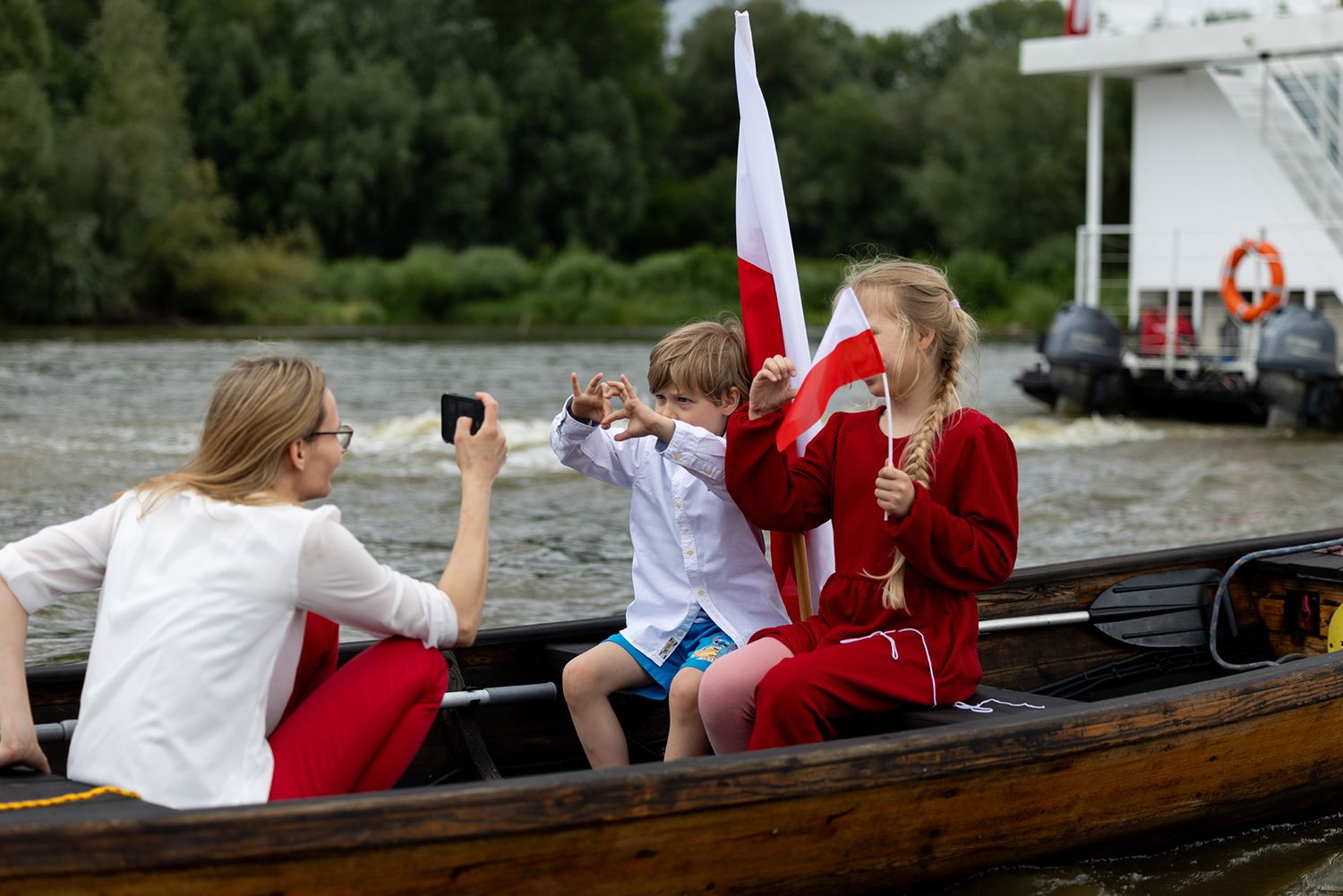 Family with Polish flag celebrating on wooden boat, woman photographing boy on river.