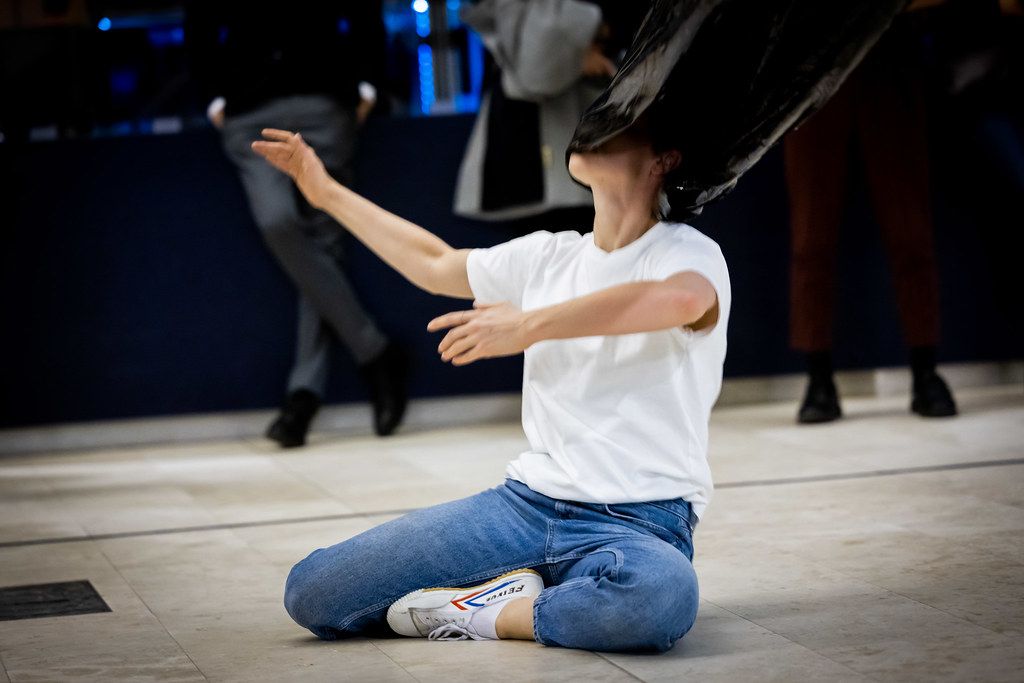 Female dancer sits on floor with arms outstretched beneath a large black object indoors.