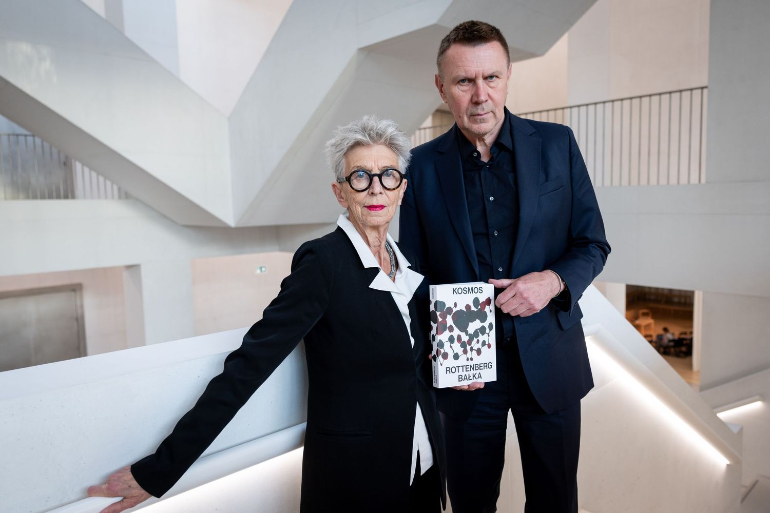 Man and woman posing with book in contemporary white building.