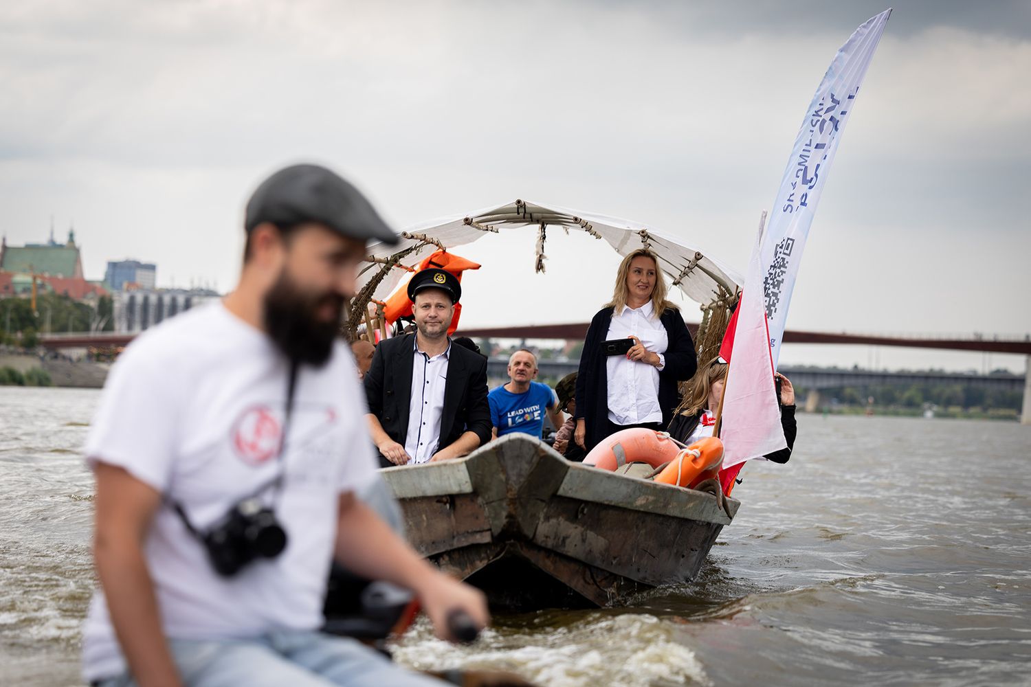 People on traditional wooden boat with white flag near city bridge on river.