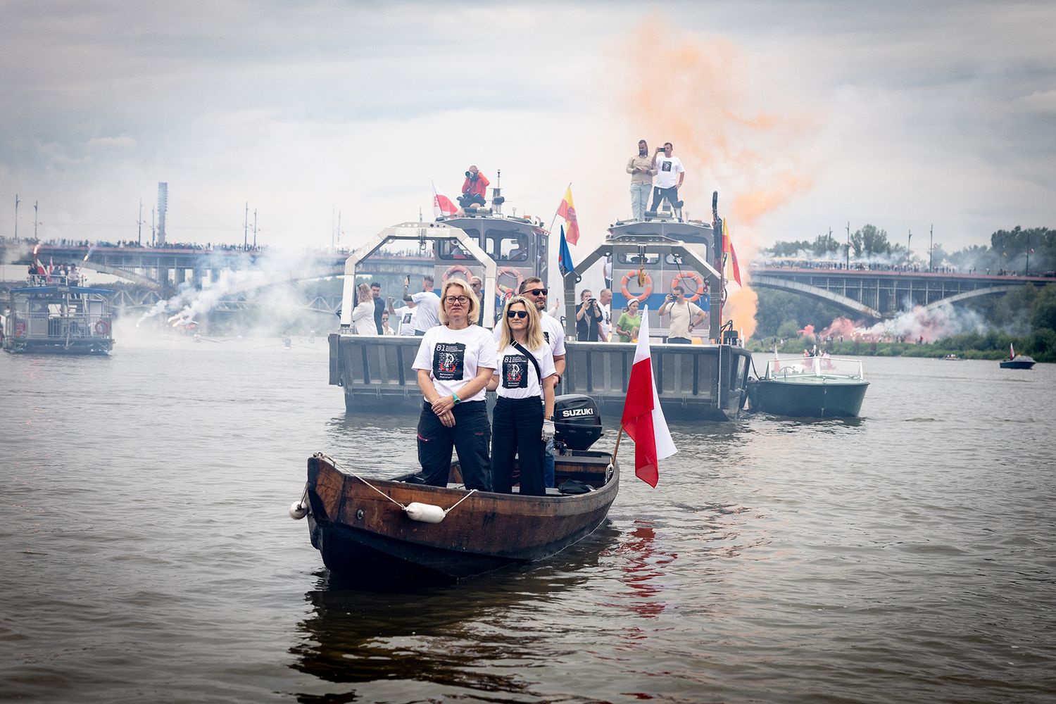 Two women in white shirts stand on boat amid smoky river flotilla.