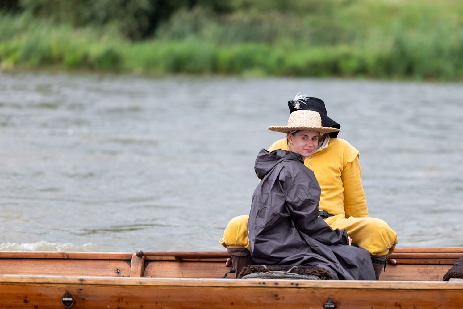 Child in yellow outfit and straw hat sitting in wooden rowboat on calm river.