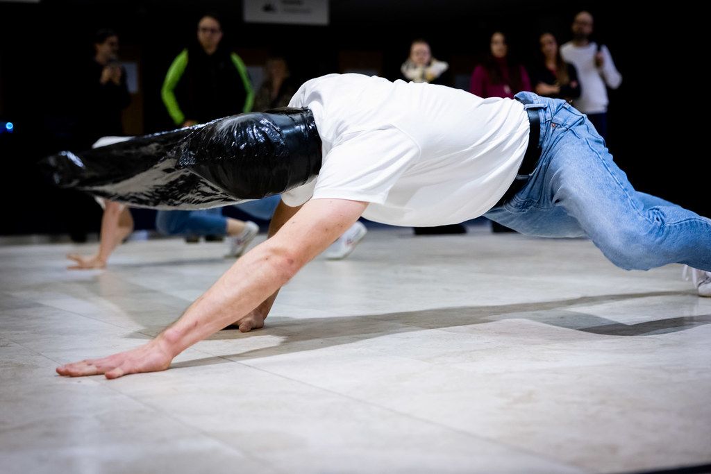 Breakdancer performs a low push-up move on a shiny floor wearing a white shirt and jeans.