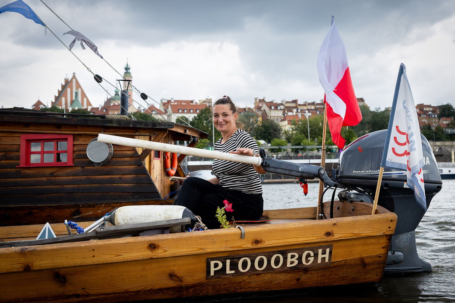 Woman posing with oar on wooden boat named PLOOOBGH with Polish flag on river.