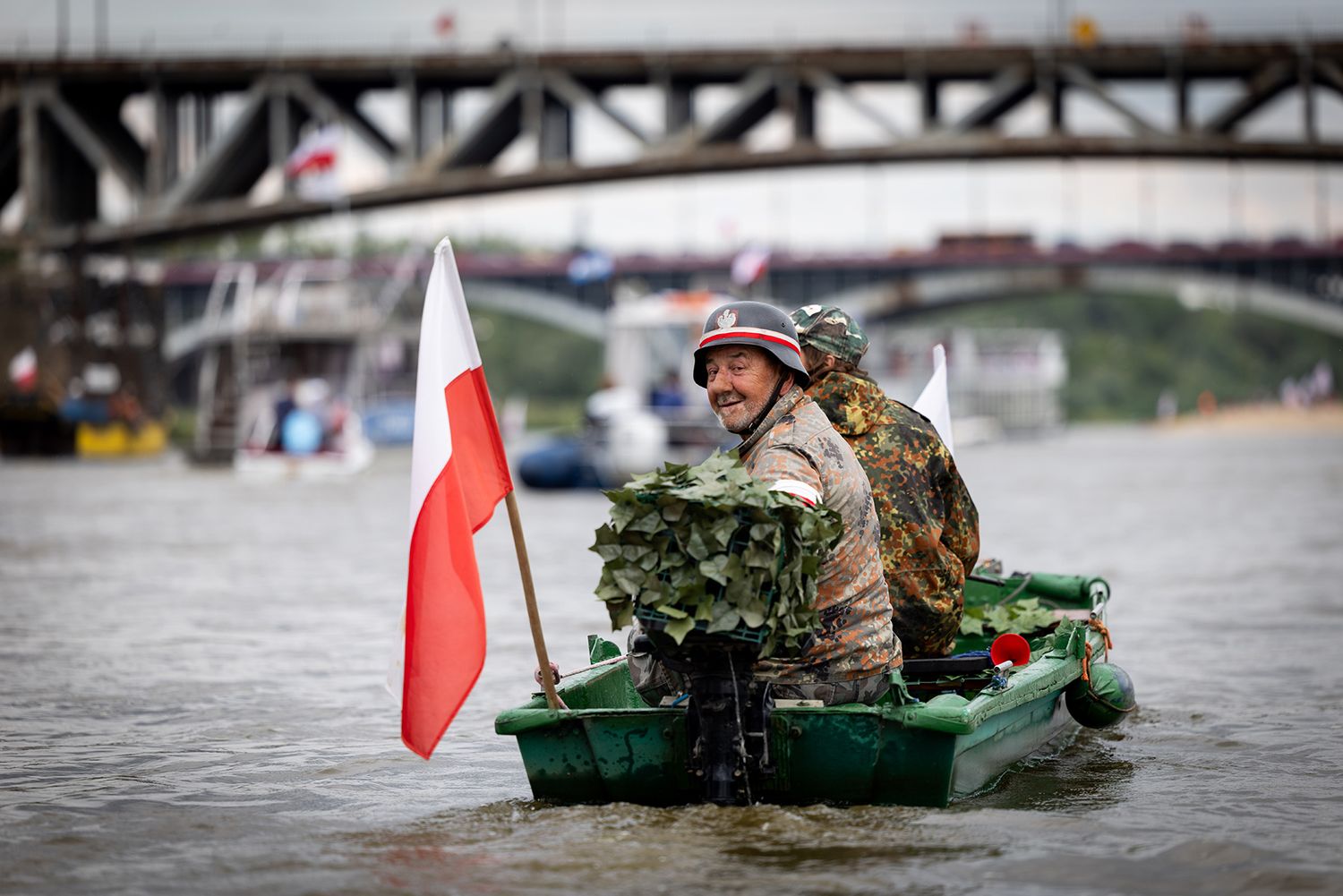 Person in camouflage on green boat with Polish flag on river.