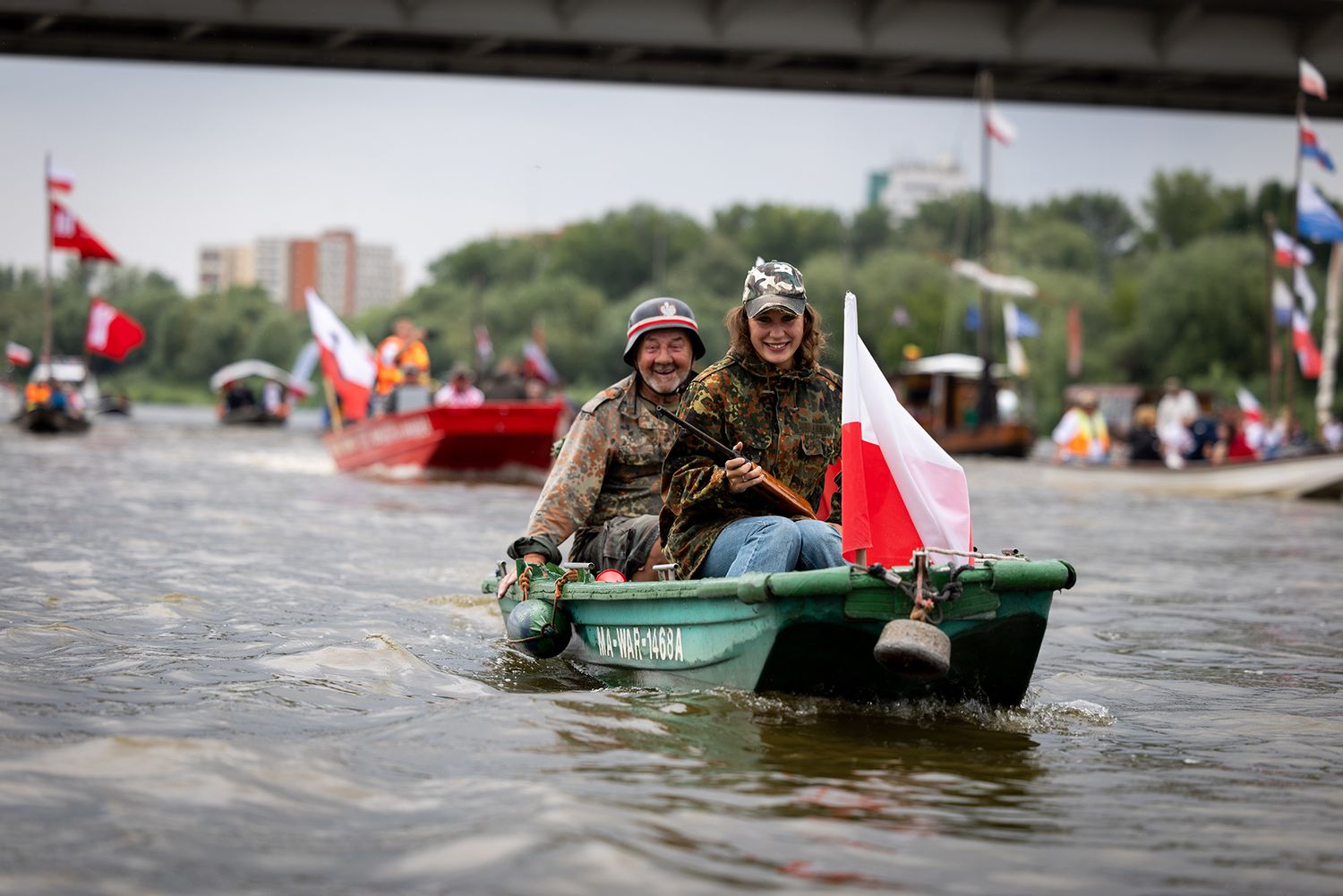 Two people in military gear on green boat with Polish flag on river.
