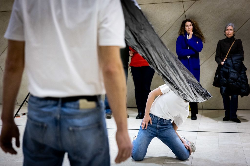 Performer kneels under a large black object while audience watches in a contemporary building.