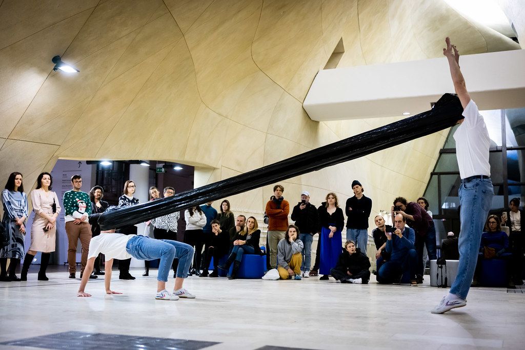 Performers execute breakdance moves under a large black pole in a modern indoor venue.