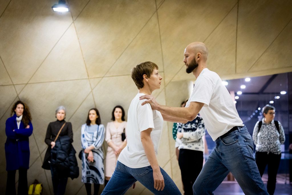 Two people in white shirts practicing self-defense moves while observers watch indoors.