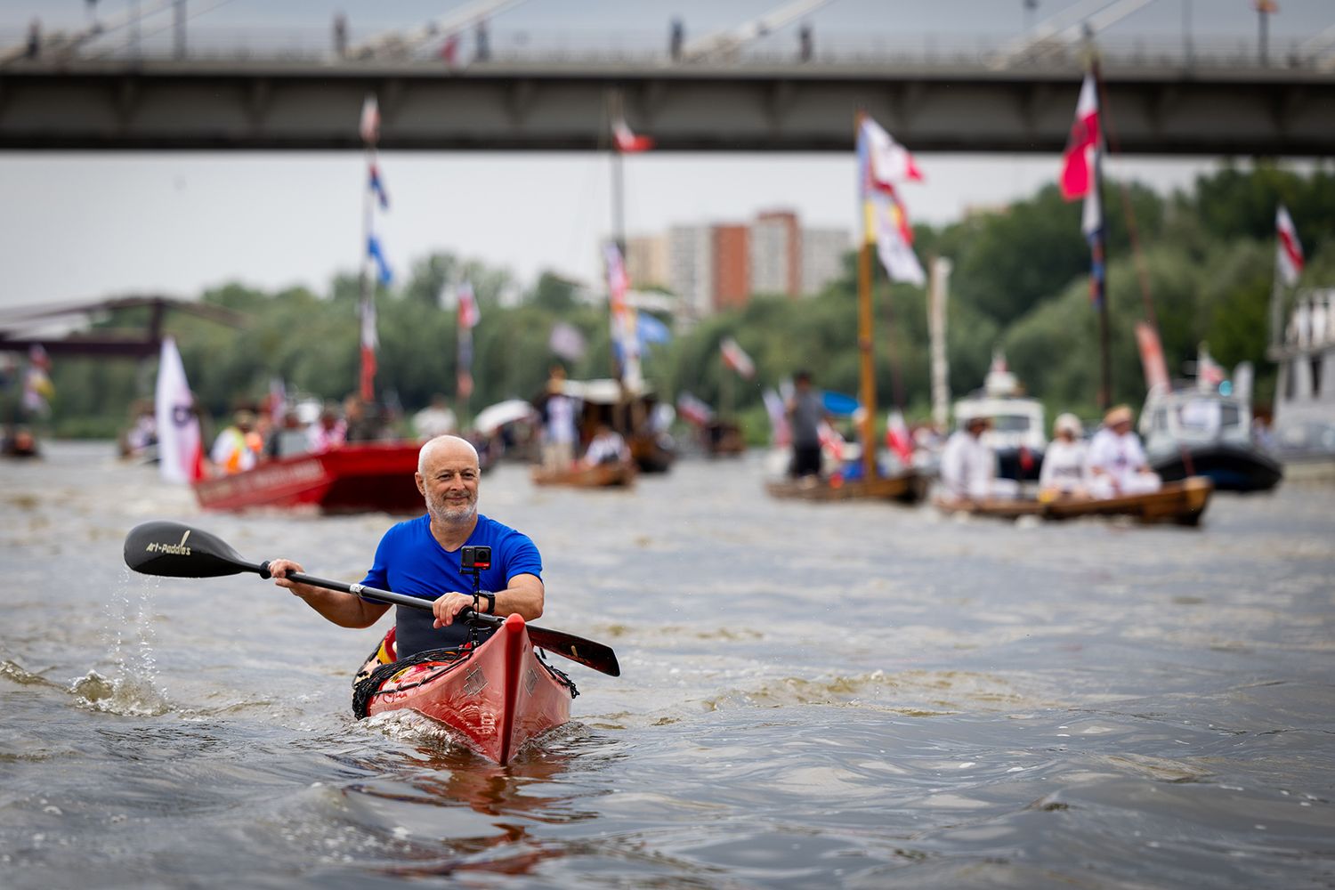 Man paddling kayak leads flotilla of boats with flags on river.