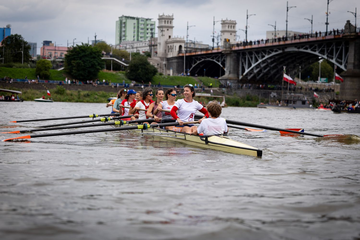 Women rowing eight-person scull on river with bridge in background.