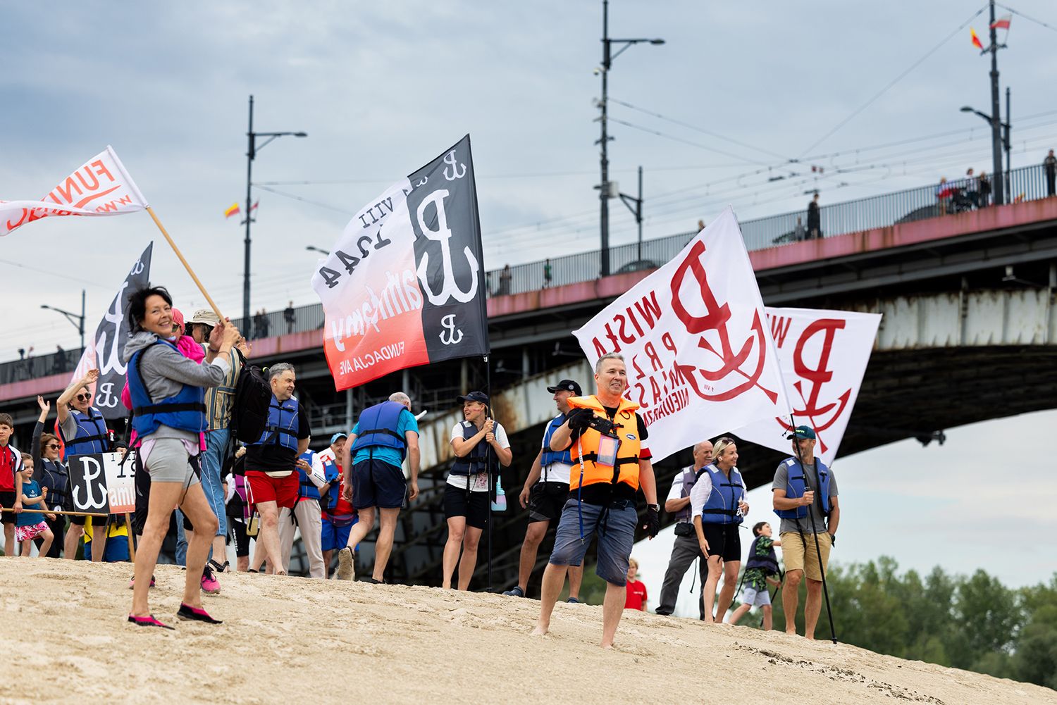 People carrying Polish uprising flags and banners walking along sandy riverbank.