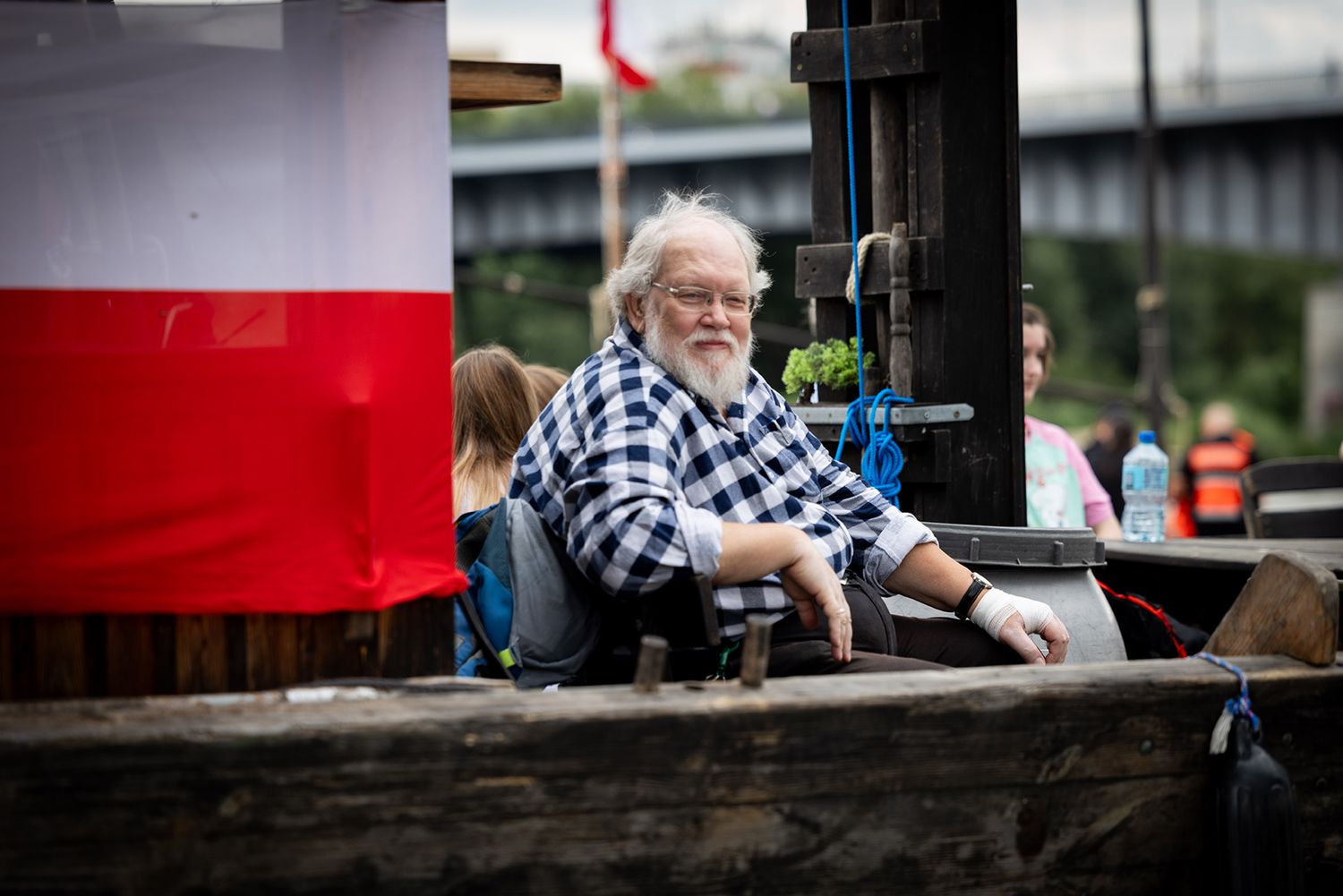 Elderly bearded man relaxing on wooden boat with Polish flag during river event.